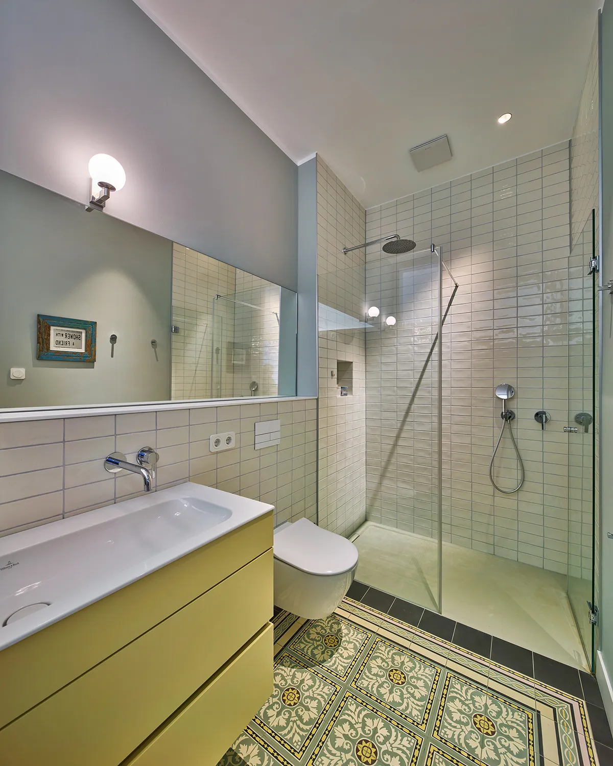 Modernisiertes Badezimmer mit bodengleicher Dusche, hellgelbem Waschtisch und historischen Jugendstilfliesen.